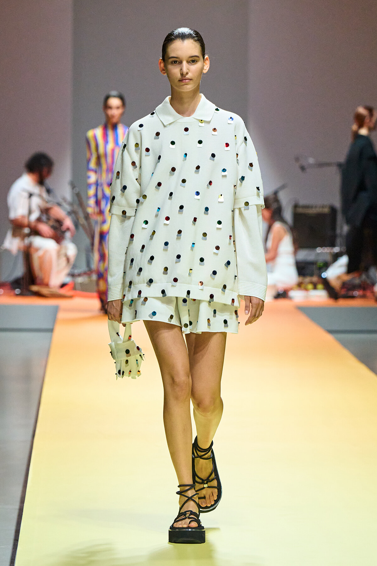 Model walks the runway in a white oversized shirt and matching shorts decorated with multicolored dots, carrying a small coordinating bag with strappy sandals.]','Model on the catwalk wearing a white polka-dot outfit (top and shorts) with a tiny bag and black sandals.