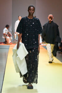 Model walks a runway in a black, oversized dress covered with circular embellishments, holding a pale green fabric while wearing platform sandals; yellow runway beneath.