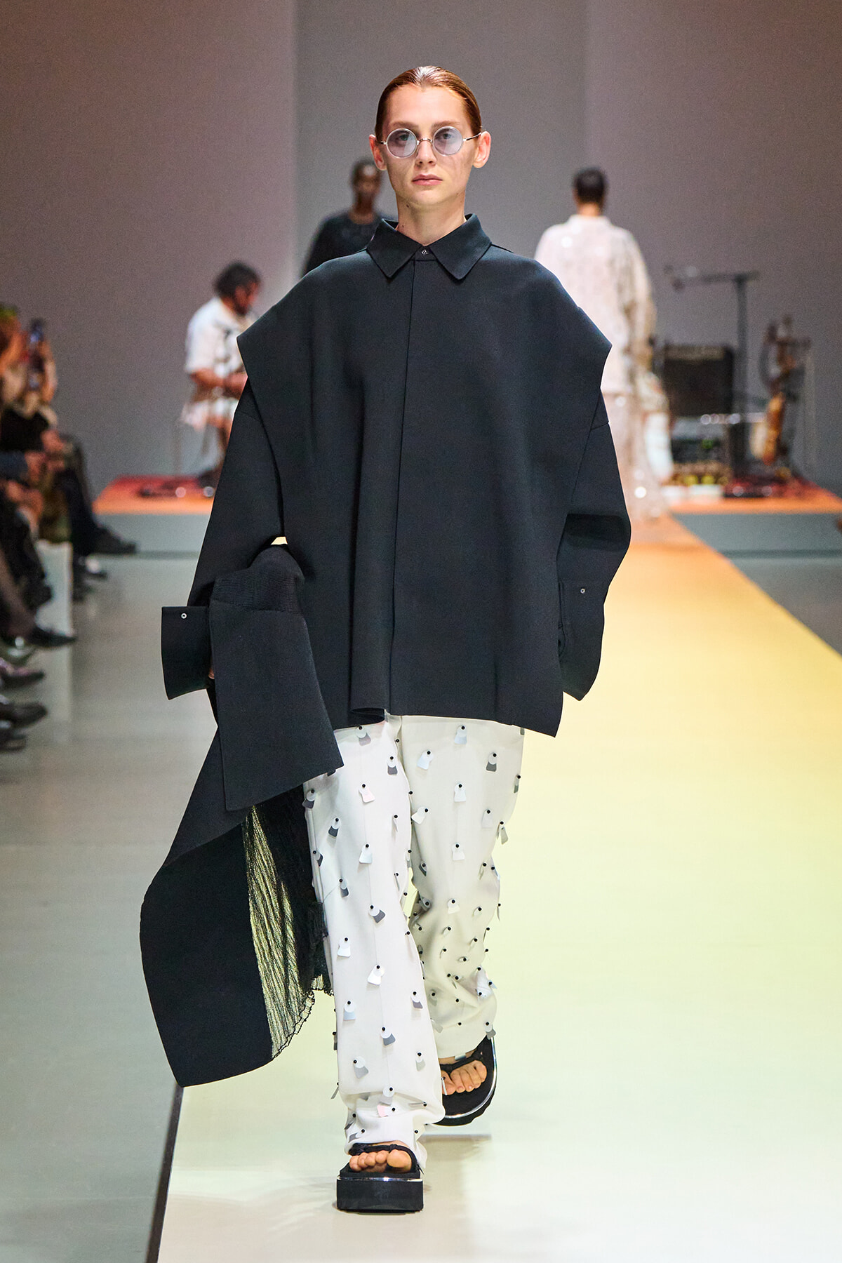 Model walks a fashion show wearing an oversized dark coat over white pants with small black shapes; round glasses and flat sandals.