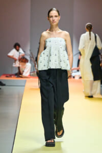 Model walks a fashion runway in a pale strapless top with small appliques and loose black trousers on a yellow catwalk.