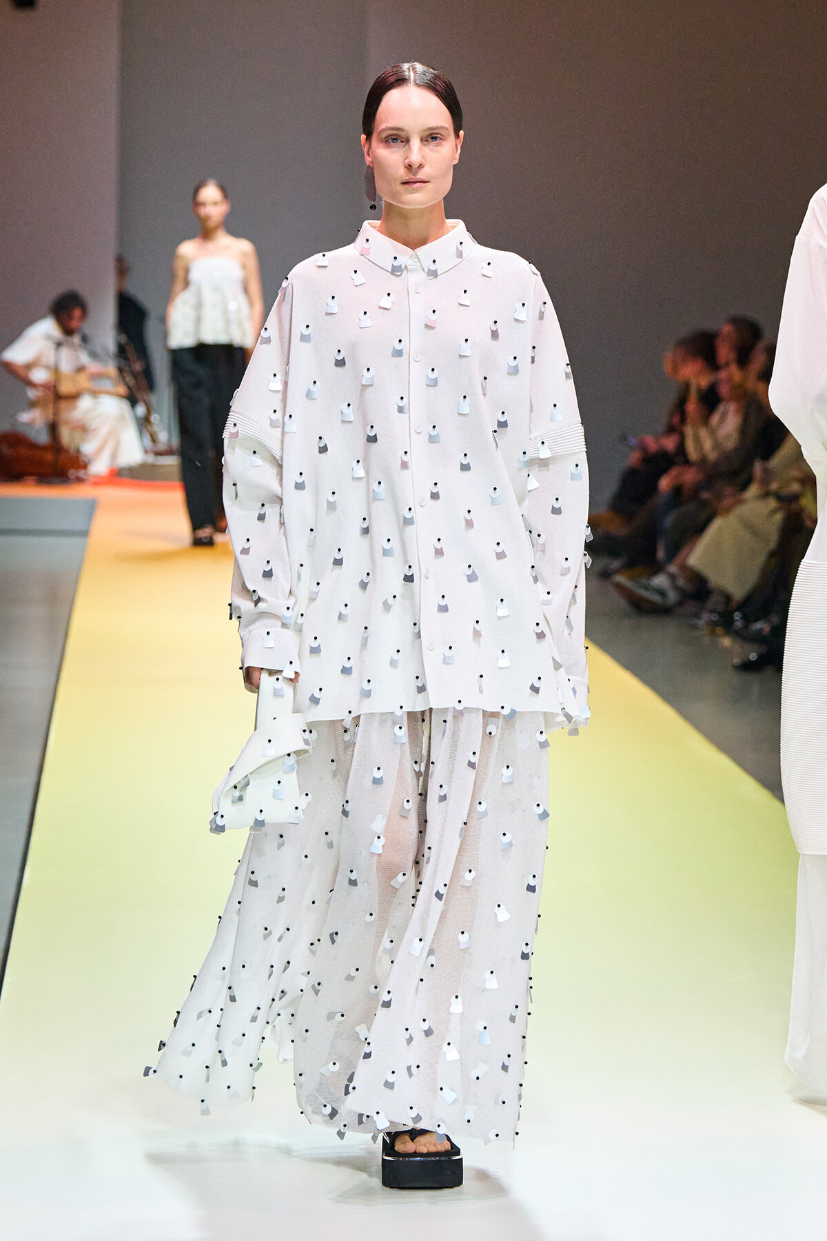 Model walks the runway in a loose white dress with small black dot appliqués and a sheer matching skirt, paired with black platform sandals.