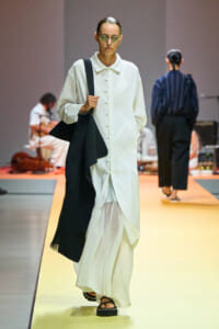 Model walking a runway in a long white button-down dress with a black draped scarf, wearing sunglasses and sandals.