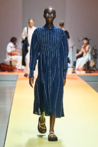 Model on a runway wearing a navy blue striped button-down midi dress with oversized sleeves, round sunglasses, and white sandals.