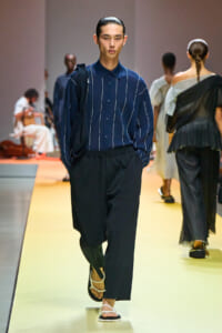 Male model walks a fashion runway in a navy striped button-down shirt, wide black pants, and white sandals, with a black shoulder bag draped over one shoulder.