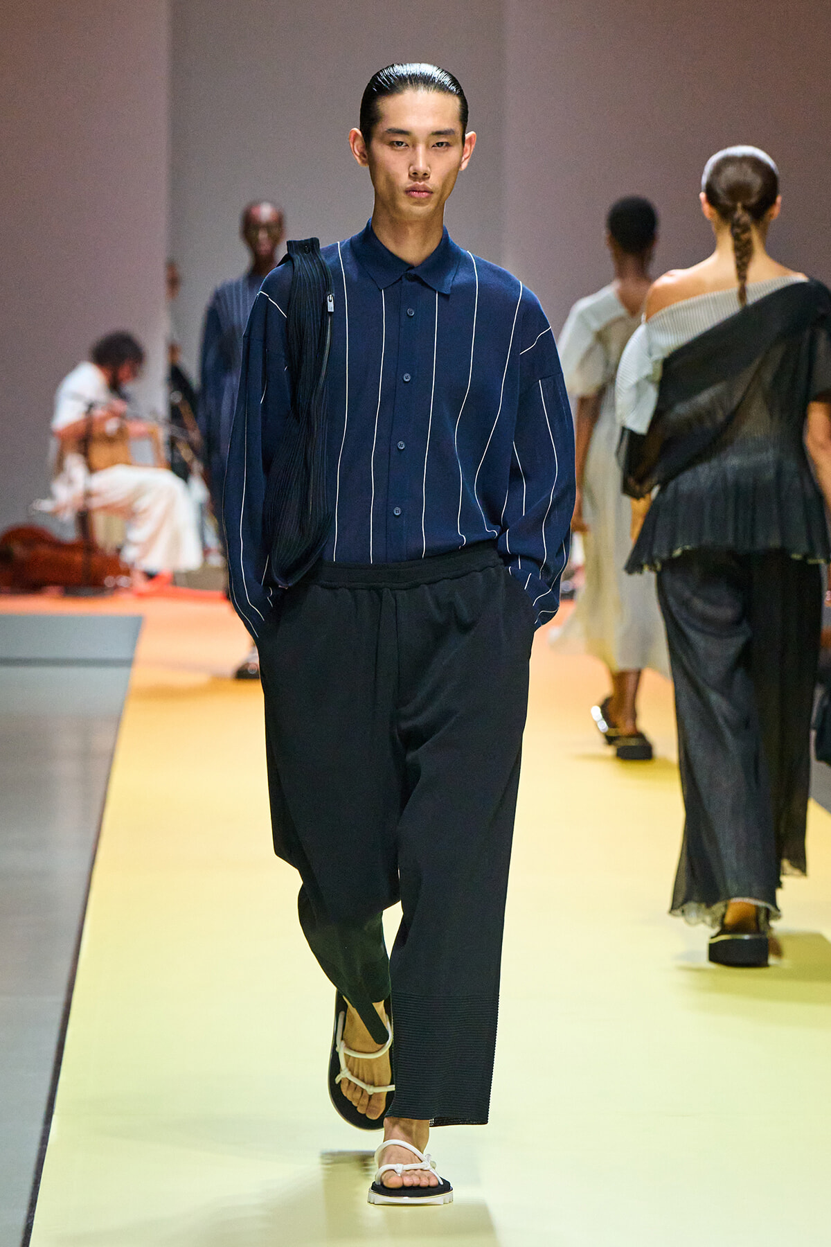 Male model walks a fashion runway in a navy striped button-down shirt, wide black pants, and white sandals, with a black shoulder bag draped over one shoulder.
