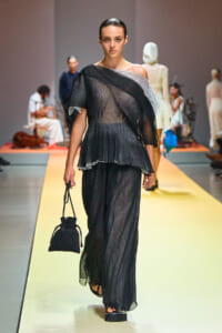 Model walks the runway in a sheer black pleated jumpsuit with an off-the-shoulder neckline, holding a small black drawstring handbag on a yellow catwalk.