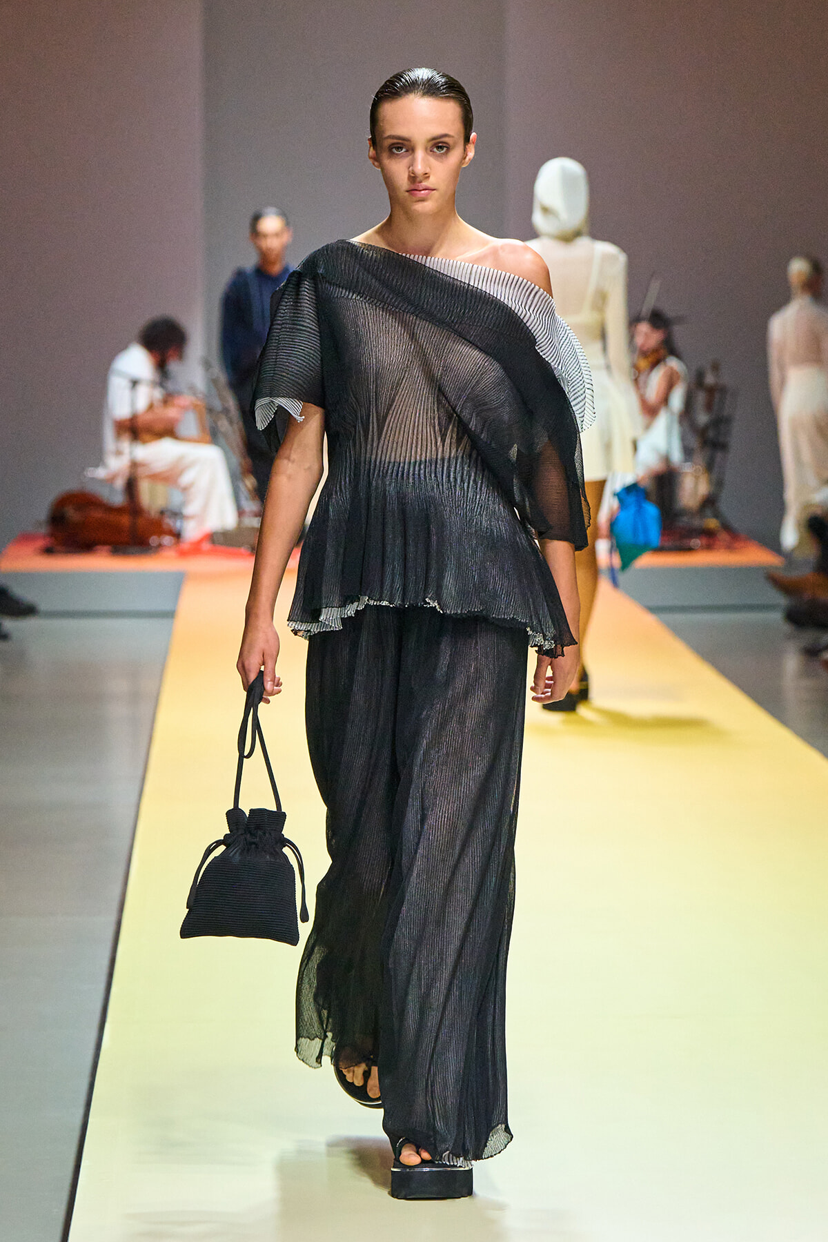 Model walks the runway in a sheer black pleated jumpsuit with an off-the-shoulder neckline, holding a small black drawstring handbag on a yellow catwalk.
