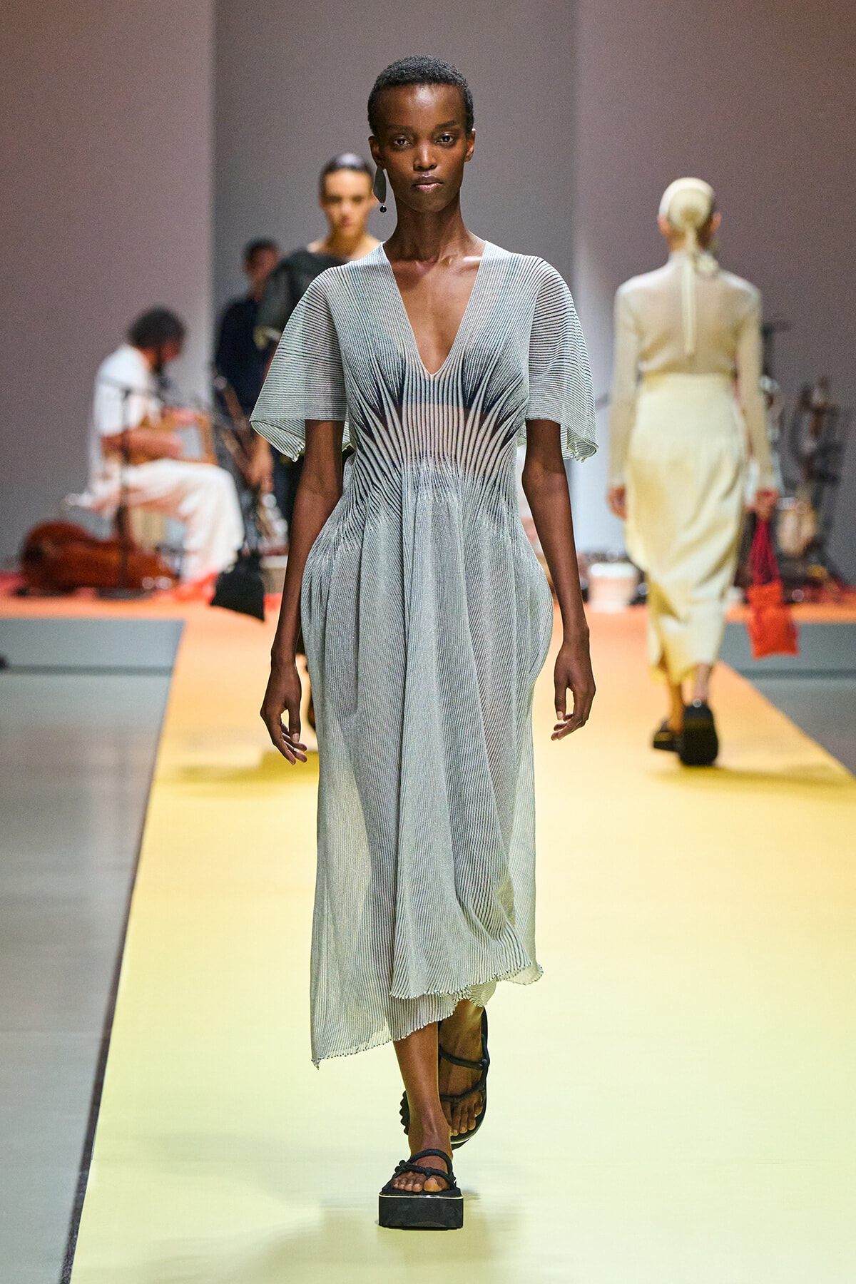 Model walking a runway in a light gray, pleated midi dress with a deep V-neck and flutter sleeves on a yellow catwalk.