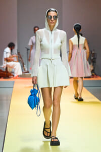 Model walking the runway in a white hooded sheer top and skirt set, wearing sunglasses and black platform sandals, carrying a bright blue bag.