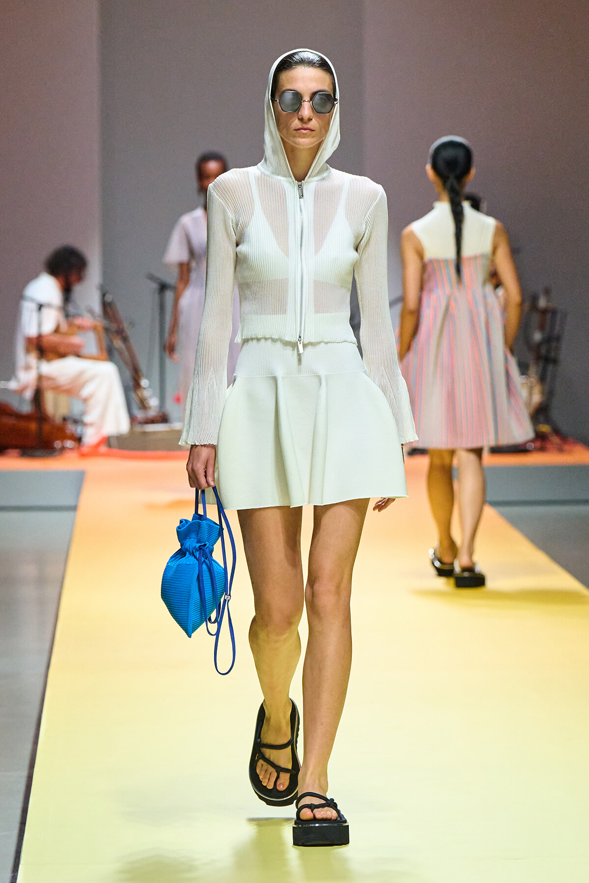 Model walking the runway in a white hooded sheer top and skirt set, wearing sunglasses and black platform sandals, carrying a bright blue bag.