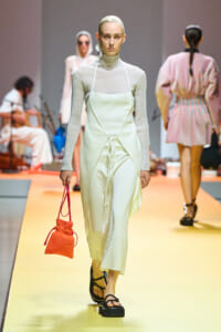 Model walking the runway in an ivory sleeveless apron over a ribbed turtleneck, carrying an orange drawstring bag.