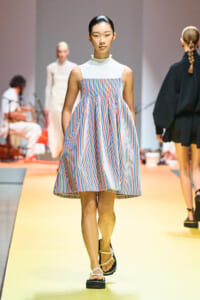 Model walking a fashion runway in a sleeveless white top and a colorful striped A-line dress, sandals on a yellow catwalk with blurred background models and crew behind her.