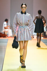 Model walking a fashion runway in a colorful, striped belted mini dress with a black collar, sunglasses, and black strappy sandals, carrying a dark knit handbag.