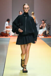Model walks a runway in a black ribbed zip-up top with a high neck and matching pleated mini skirt, carrying a black bag; yellow catwalk and blurred audience in background.