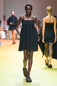 Front view of a dark-skinned model walking a fashion show runway in a black pleated spaghetti-strap dress and strappy sandals, with other models in the background.