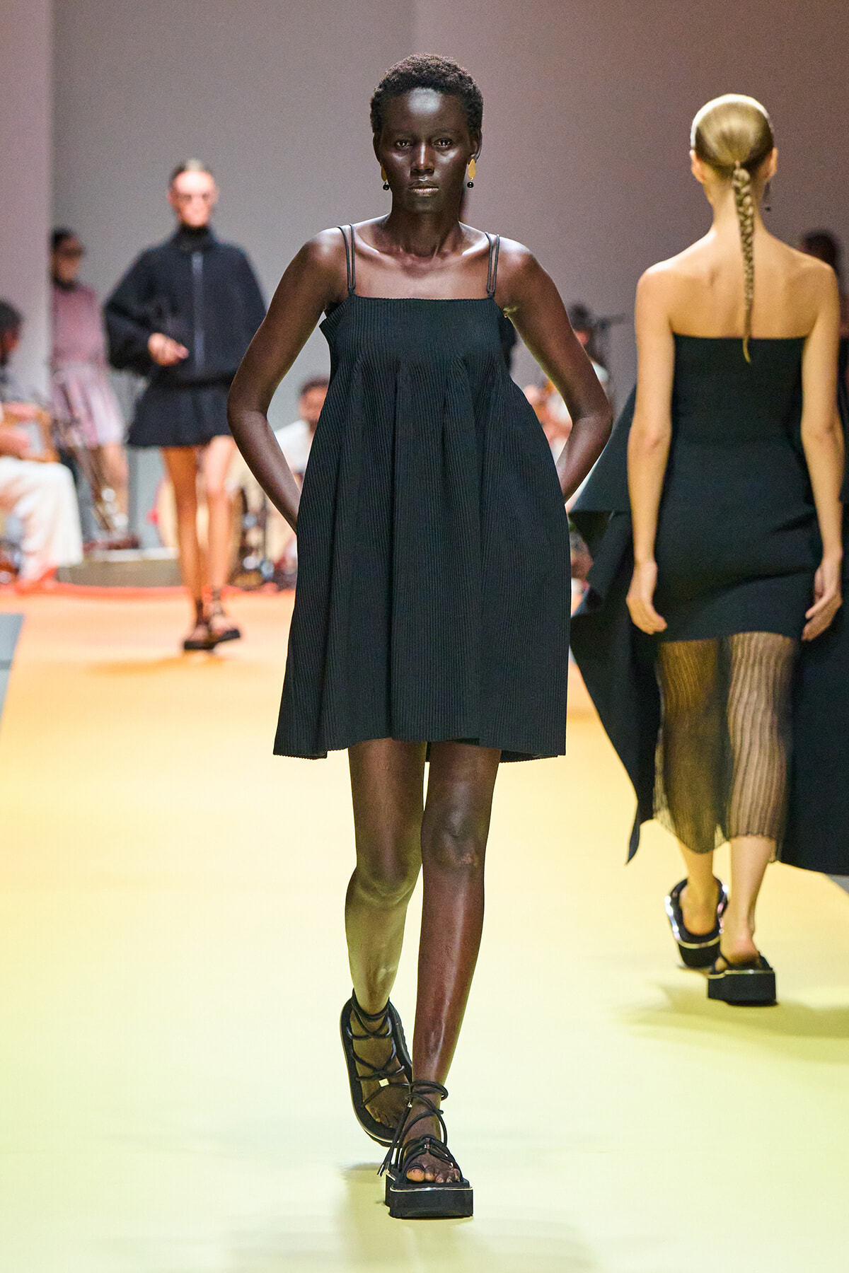 Front view of a dark-skinned model walking a fashion show runway in a black pleated spaghetti-strap dress and strappy sandals, with other models in the background.
