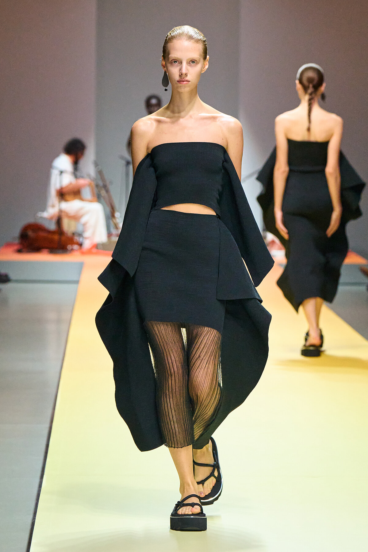 Model walking a runway in a black off-the-shoulder crop top and matching asymmetrical cape-style skirt with sheer striped tights and sandals.