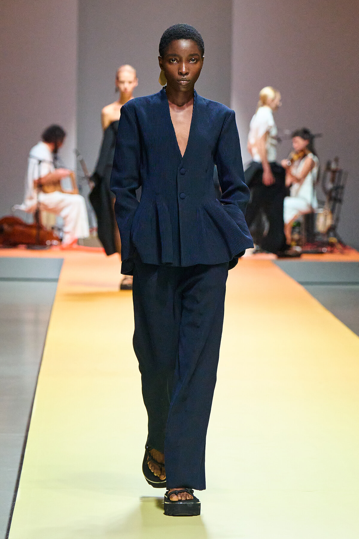 Model walks a fashion runway in a navy blue pleated blazer and matching wide-leg pants, yellow catwalk behind her, musicians in the background.