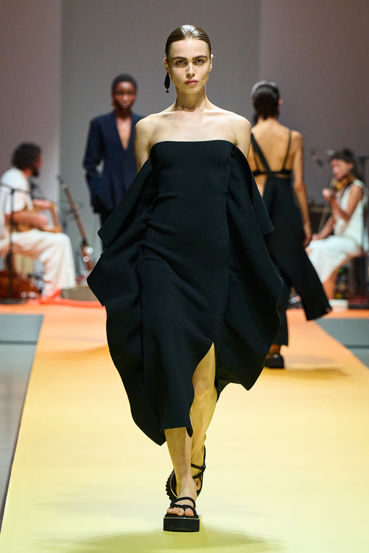 Model in a black off-shoulder dress with dramatic cape-like sleeves walking a yellow runway; focused expression, minimal accessories.