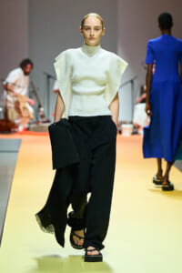 Model walks the runway in a white high-neck top with exaggerated cape-like sleeves and black wide-leg trousers, holding a black shawl, on a yellow catwalk surface.
