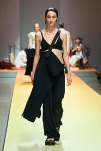 Model walks the runway in a black, asymmetrical two-piece outfit with a diagonal crossbody strap and a draped, wide-leg skirt; platform sandals visible.