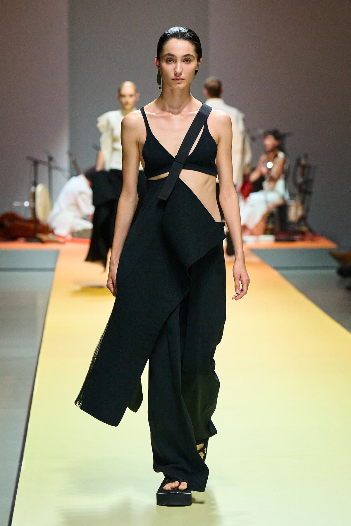 Model walks the runway in a black, asymmetrical two-piece outfit with a diagonal crossbody strap and a draped, wide-leg skirt; platform sandals visible.