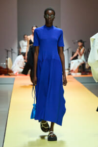 Model walks a runway in a cobalt blue short-sleeve midi dress, round sunglasses, white sandals, and a small blue bag.