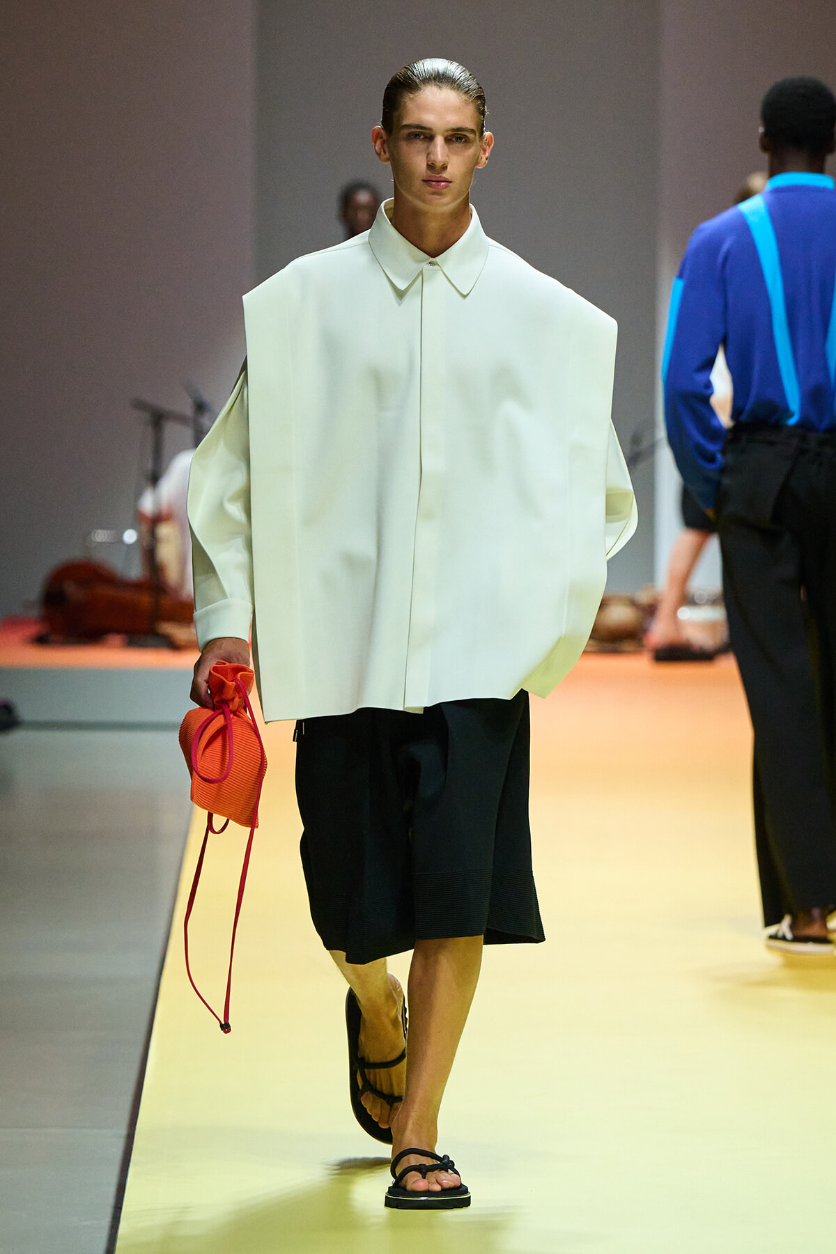Model walks a fashion runway wearing an oversized pale shirt, knee-length black culottes, and black sandals, carrying an orange bag.