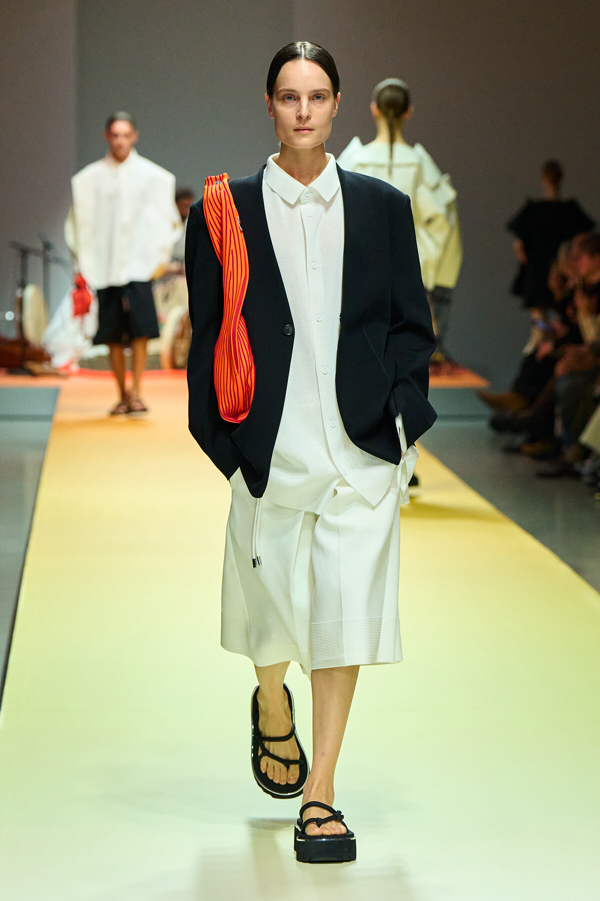 Model on a runway wearing a white shirt dress with a black blazer, carrying a bright red striped clutch.