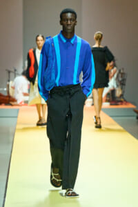 Male model walking a fashion runway in a cobalt blue shirt with bright blue stripes and loose black pants, sandals visible at the front.