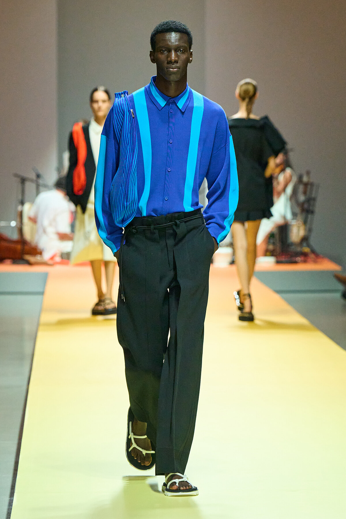 Male model walking a fashion runway in a cobalt blue shirt with bright blue stripes and loose black pants, sandals visible at the front.