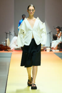 Model walks a fashion runway in a white oversized blazer with a deep V and black wide shorts, wearing glasses and black platform sandals on a yellow catwalk.