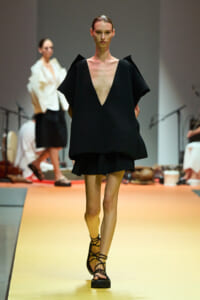 Model on a runway wearing an oversized black dress with a deep V neckline and cape-like sleeves, paired with strappy sandals.