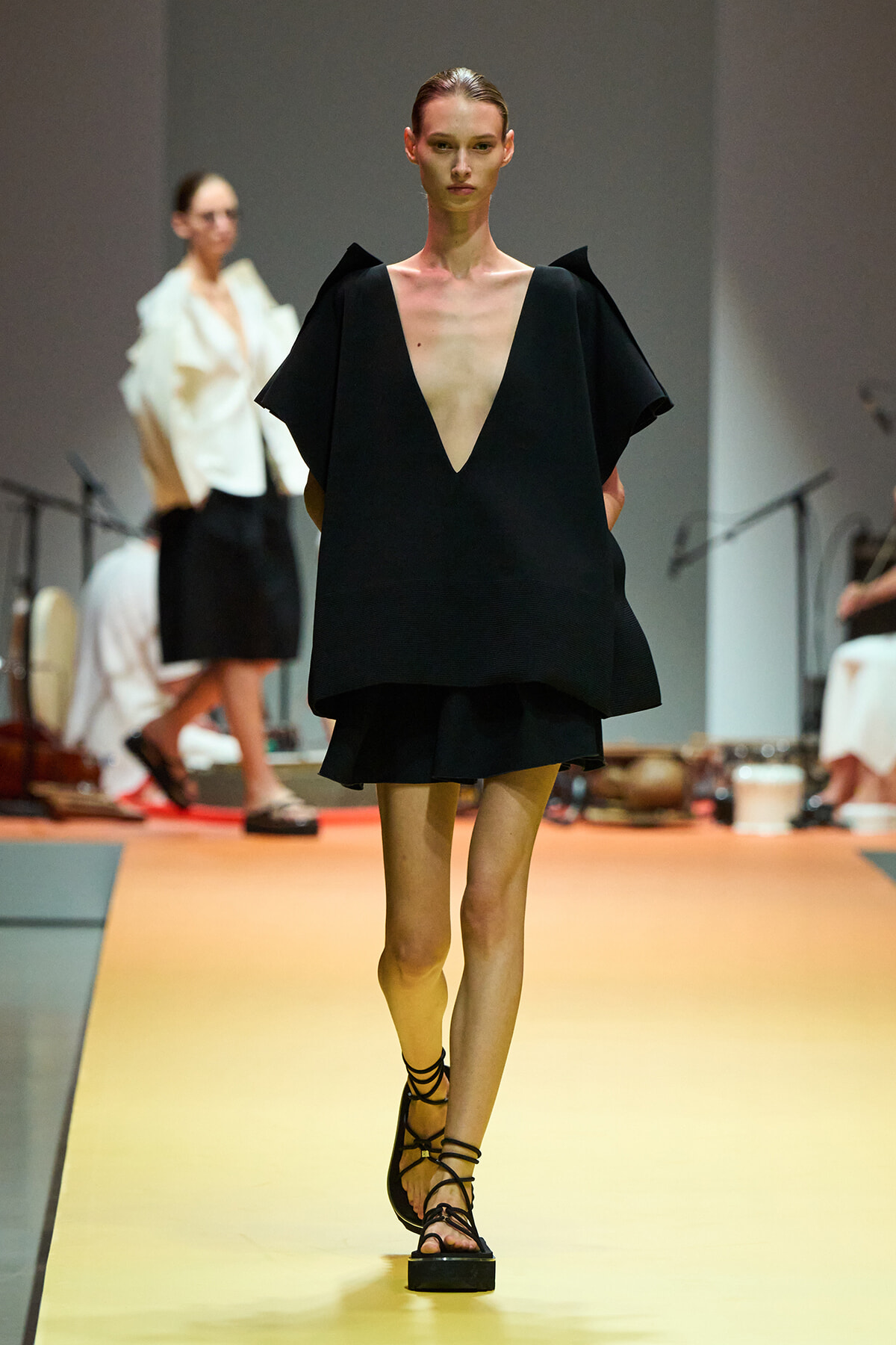Model on a runway wearing an oversized black dress with a deep V neckline and cape-like sleeves, paired with strappy sandals.