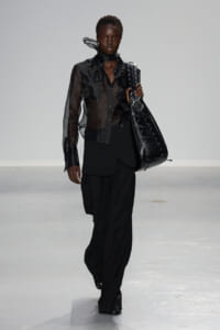 Model walks the runway in a sheer black blouse and oversized black pants, carrying a large black lace-up tote bag.