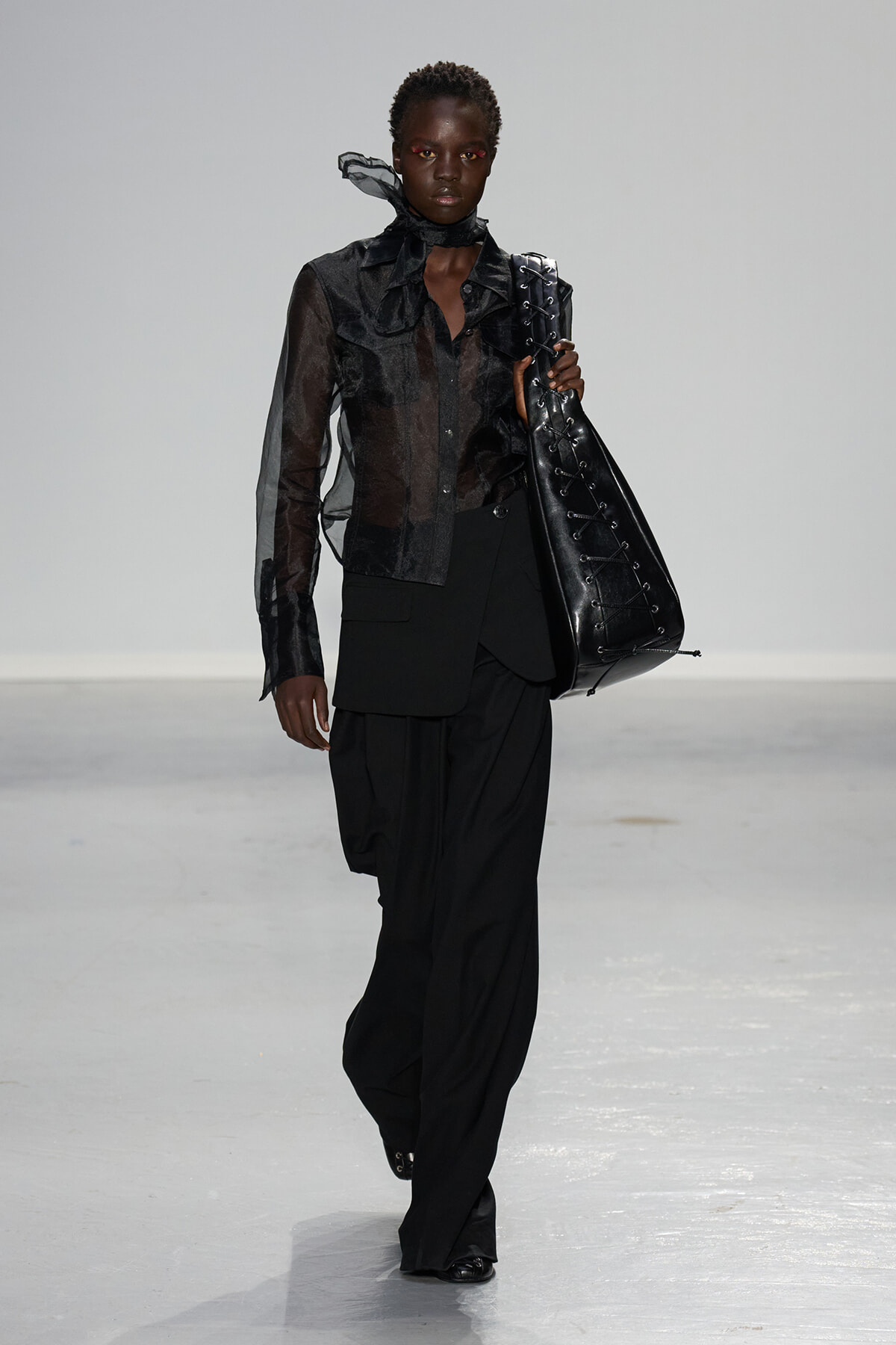 Model walks the runway in a sheer black blouse and oversized black pants, carrying a large black lace-up tote bag.