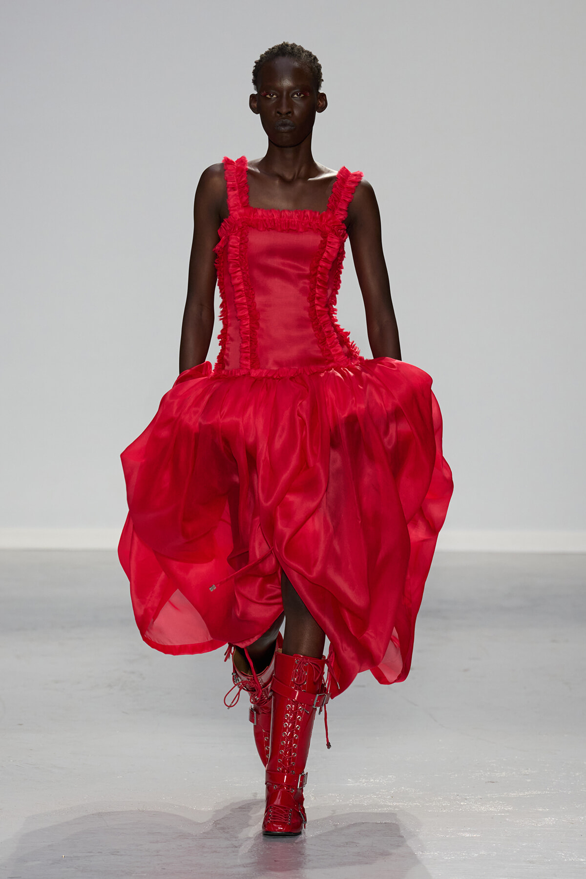 Model on a runway wearing a bright red satin dress with frilled trim and a voluminous, draped skirt, paired with red lace-up boots.