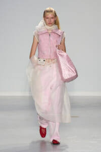 Runway model in a pink cropped vest, sheer scarf, and voluminous pale pink skirt, carrying a pink studded tote.