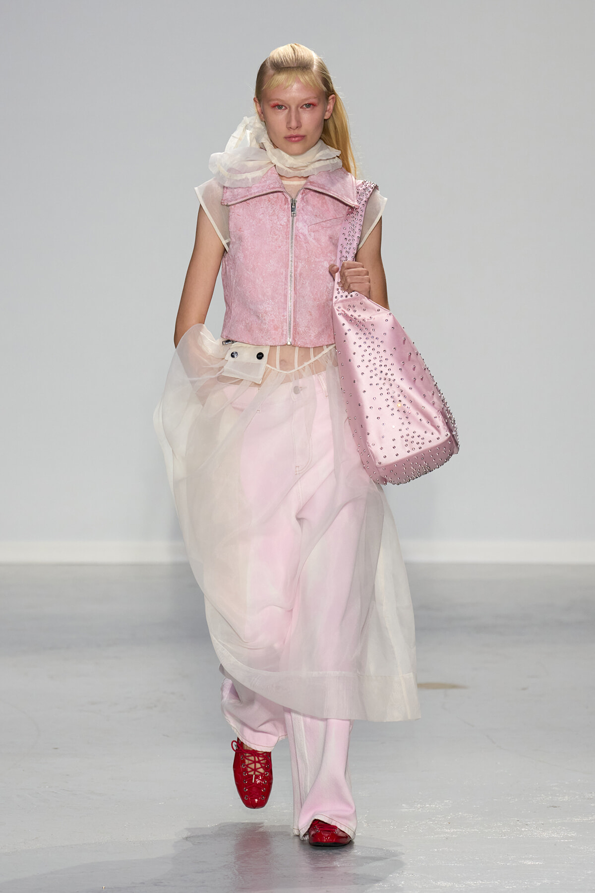 Runway model in a pink cropped vest, sheer scarf, and voluminous pale pink skirt, carrying a pink studded tote.