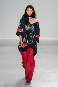 Model walks the runway in an oversized black graphic tunic with white lettering and bright red wide-leg pants, scarf at neck.