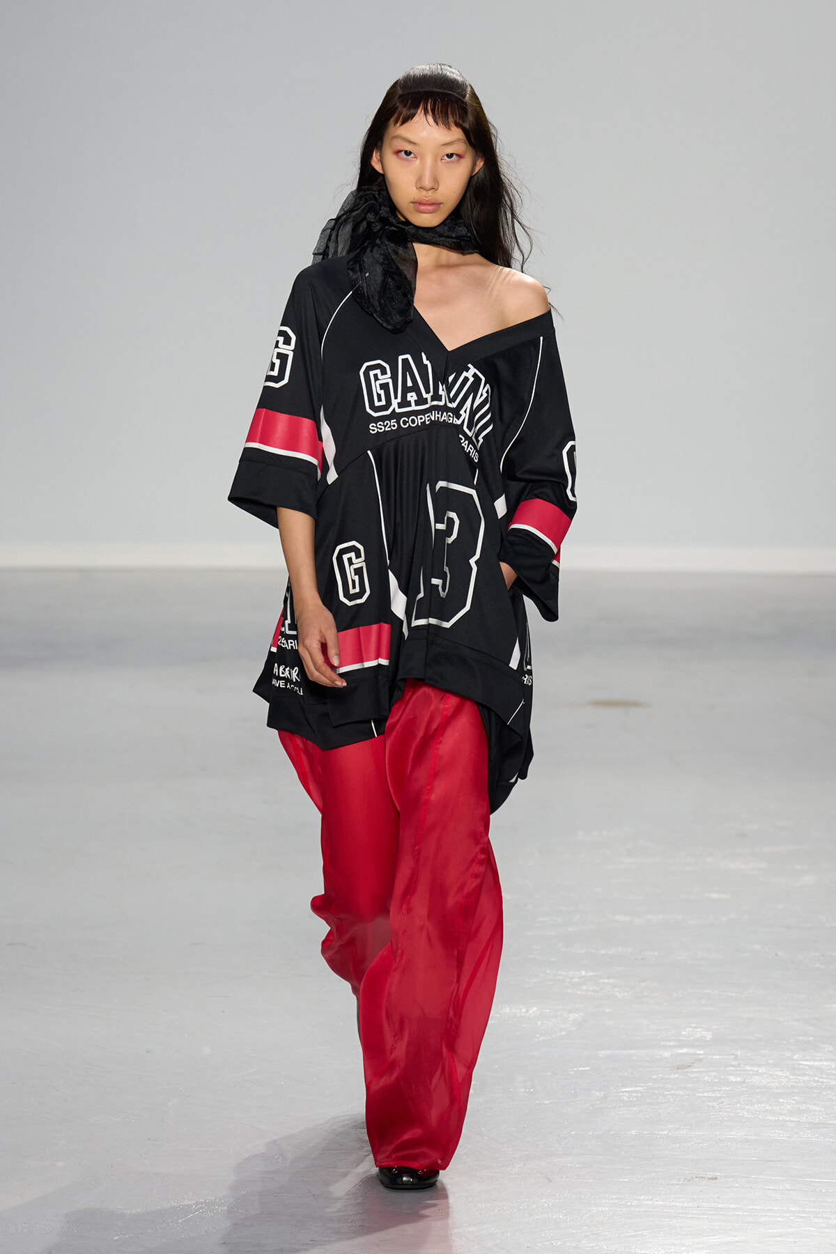 Model walks the runway in an oversized black graphic tunic with white lettering and bright red wide-leg pants, scarf at neck.