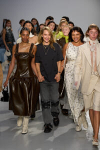 Designer (center) walks hand-in-hand with models at a fashion show, surrounded by a line of diverse models in stylish outfits.