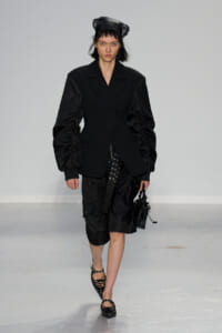 Model on a runway wearing an all-black ensemble: oversized, structured blazer with dramatic ruched sleeves, paired with a black skirt/pants and strappy flats; black headscarf and small black handbag in hand.