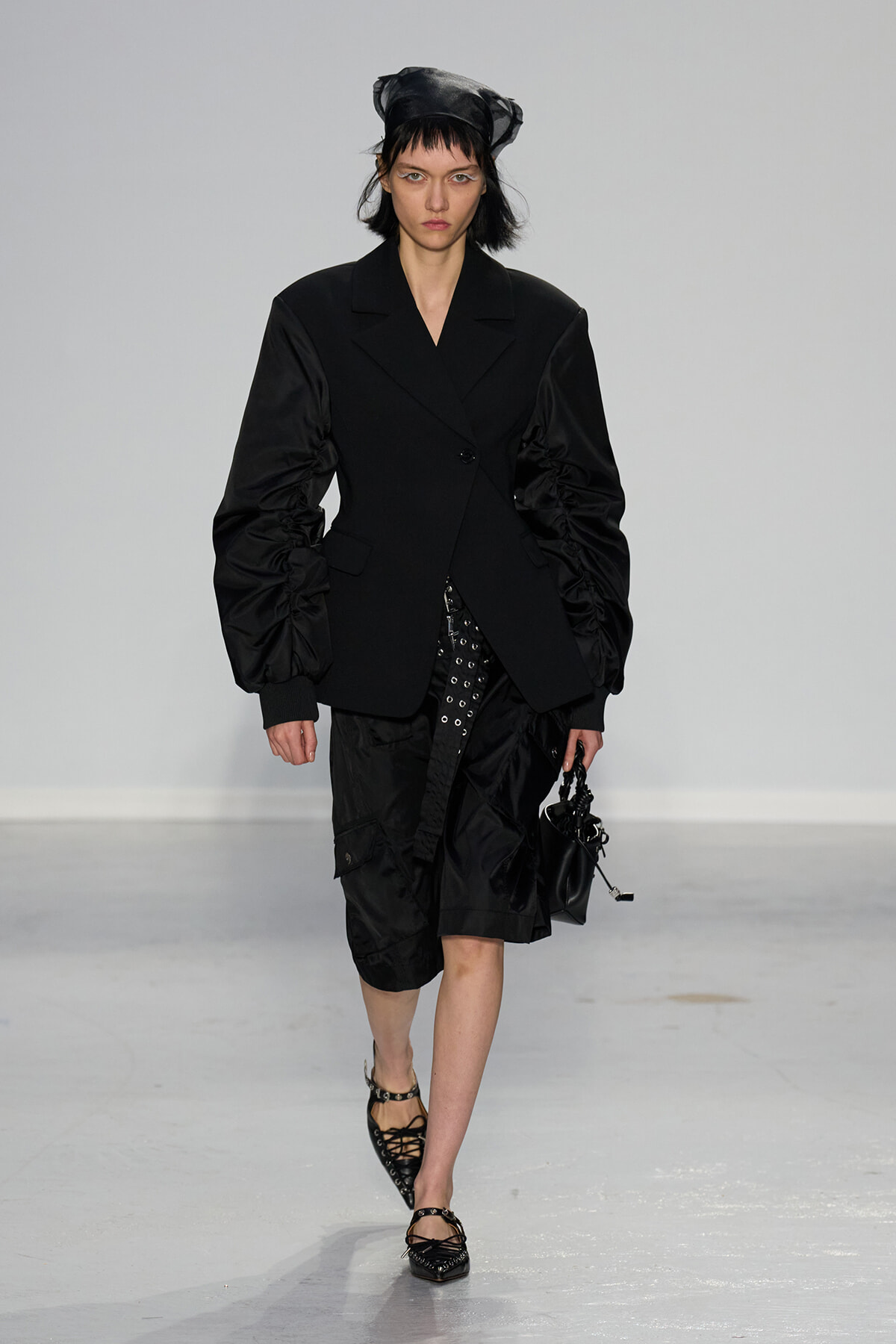 Model on a runway wearing an all-black ensemble: oversized, structured blazer with dramatic ruched sleeves, paired with a black skirt/pants and strappy flats; black headscarf and small black handbag in hand.