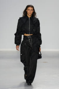 Female model walking a runway in an all-black cropped utility jacket and wide cargo pants with silver buckles and hardware.