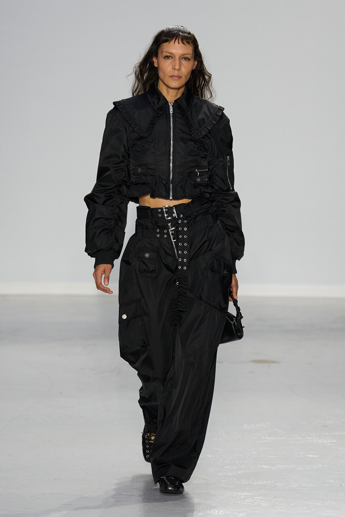 Female model walking a runway in an all-black cropped utility jacket and wide cargo pants with silver buckles and hardware.