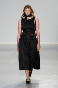Runway model in an all-black outfit: ruched sleeveless top, voluminous midi skirt with pockets, and a black neck scarf, walking forward on the runway.