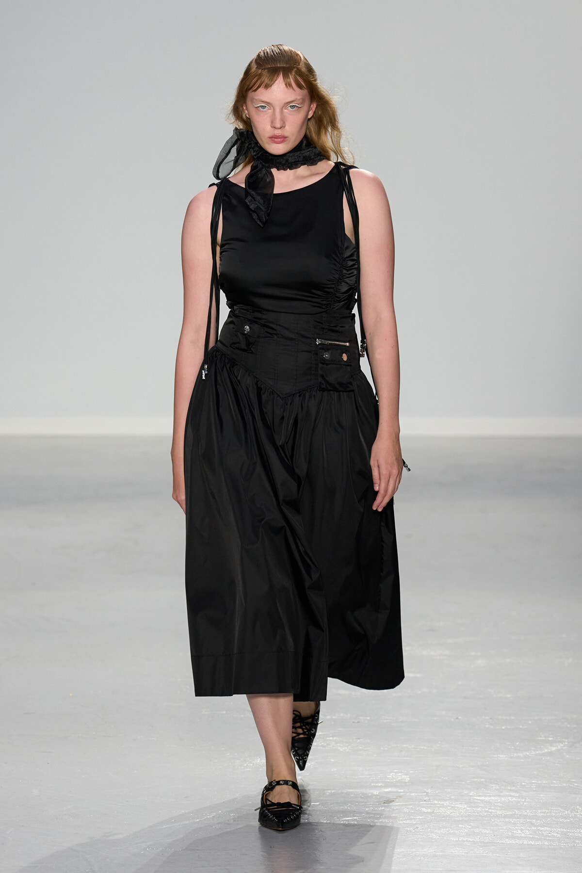 Runway model in an all-black outfit: ruched sleeveless top, voluminous midi skirt with pockets, and a black neck scarf, walking forward on the runway.