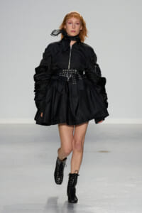 Model walking a runway in a black puff-sleeve dress with a wide studded belt and lace-up ankle boots