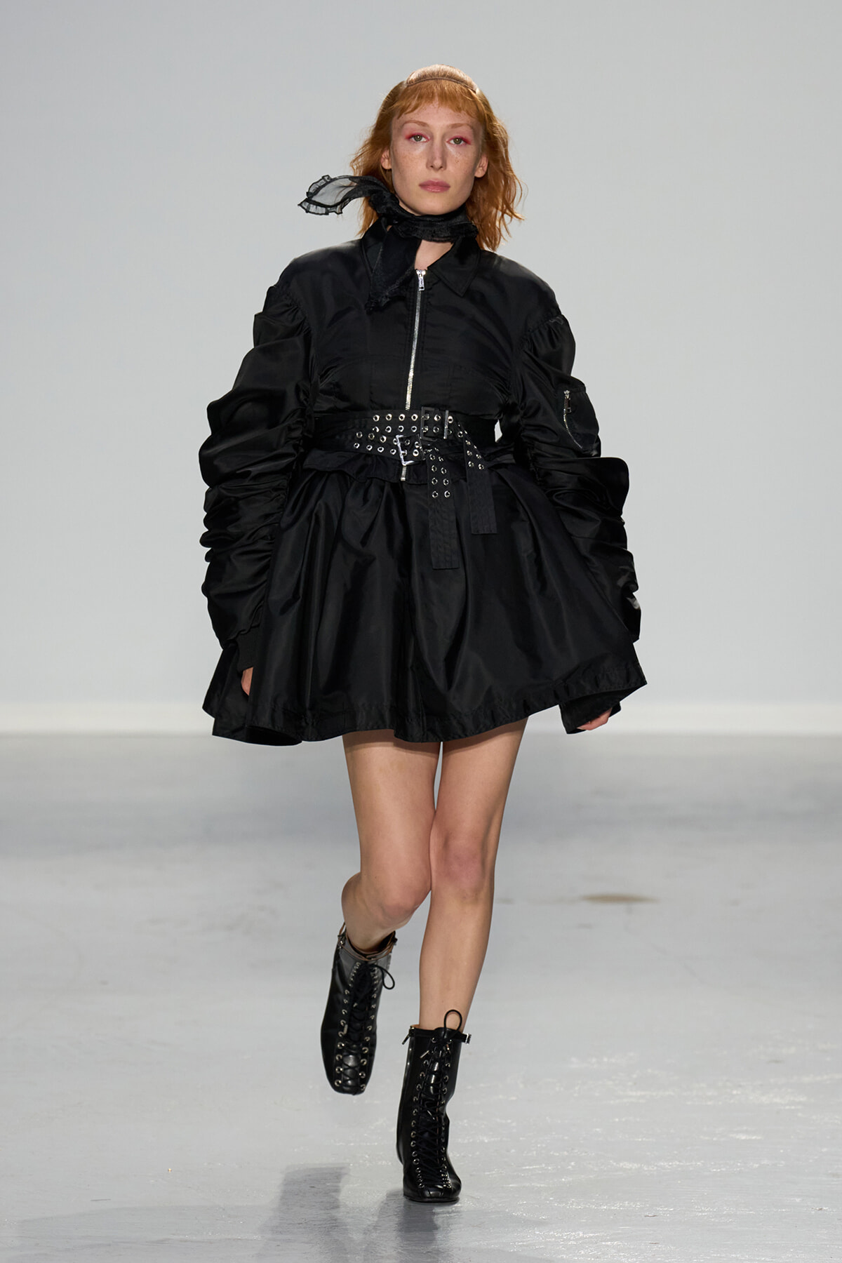 Model walking a runway in a black puff-sleeve dress with a wide studded belt and lace-up ankle boots